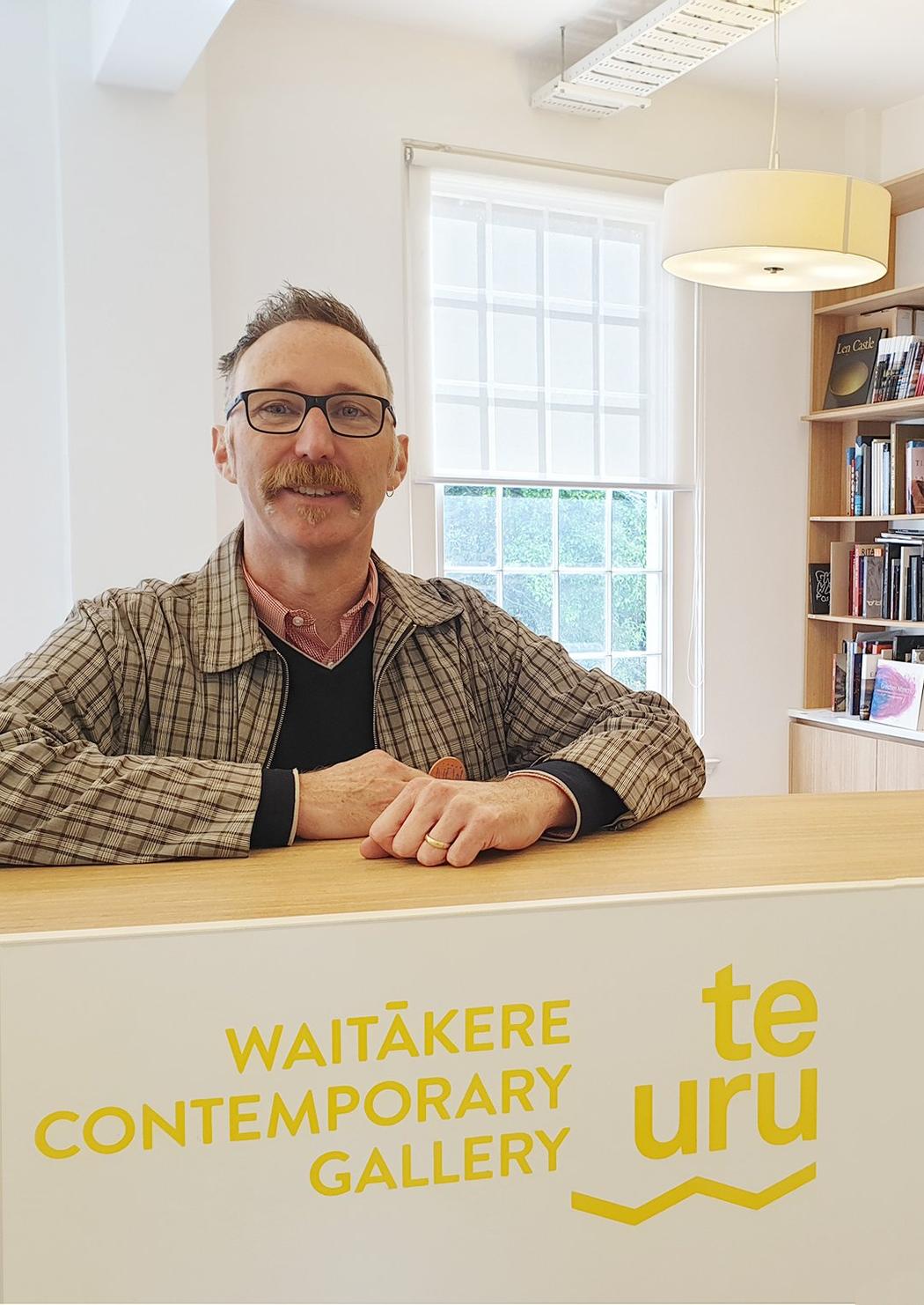 A person wearing glasses, standing behind a counter that says Te Uru Waitākere Contemporary Gallery